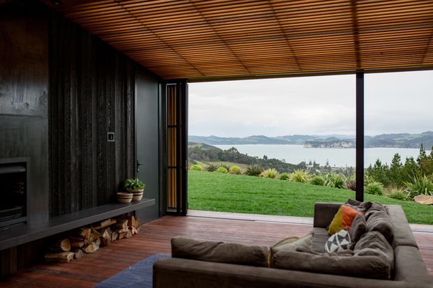 A dark, timber-lined living room opens through wide floor-to-ceiling sliding doors onto a timber deck and lawn, with expansive views across a bay and surrounding hills. The room features a low built-in fireplace wall and firewood storage, a large sectional sofa, warm timber slatted ceiling, and strong indoor–outdoor connection.