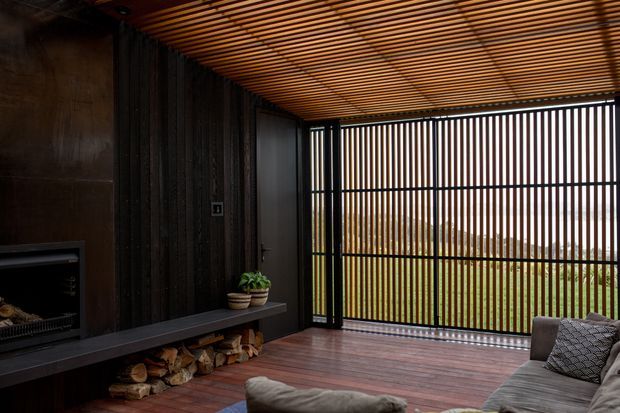 A dark, refined outdoor living room or covered patio with a linear timber-slat ceiling and matching vertical slatted screening. The space features dark timber wall cladding, a built-in fireplace with a long hearth and firewood storage beneath, warm-toned timber decking, and upholstered lounge seating. The slatted enclosure opens to a green landscape beyond, creating a sheltered indoor-outdoor entertaining area.
