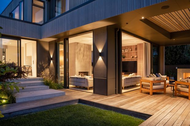 Evening exterior view of a contemporary multi-level home opening onto a timber-decked outdoor living area. Dark vertical-clad walls, extensive floor-to-ceiling glazing, warm interior lighting, landscaped steps, and a covered terrace with outdoor seating are visible. The interior living room can be seen through the open glazing.