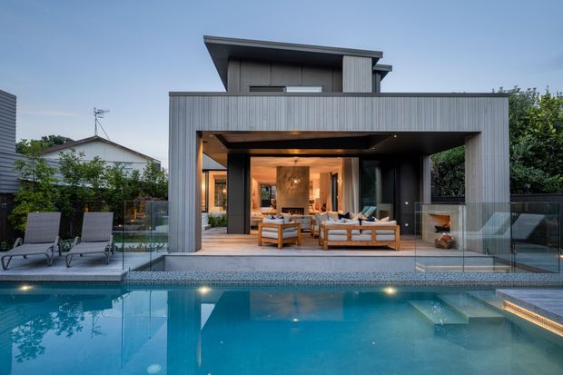 Contemporary two-storey residential house with a strongly rectilinear form, vertical grey cladding, and a large open-plan indoor-outdoor living area facing a swimming pool. The covered terrace contains lounge seating and opens directly to the pool deck, with extensive glazing and warm interior lighting. A neighbouring house and garden vegetation are visible in the background.