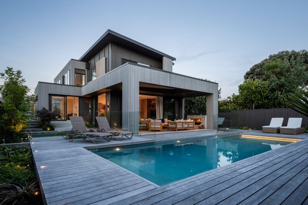Contemporary multi-level residence with dark vertical cladding, extensive glazing, and broad roof overhangs, viewed from a landscaped pool area. A timber-look deck surrounds the rectangular swimming pool, with outdoor loungers, a covered entertaining area, dining furniture, and integrated landscape planting.
