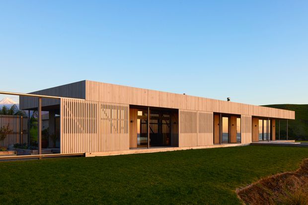 Single-storey contemporary residence set in an open landscaped site, with a long low-profile form, flat roof, extensive glazing, and warm vertical timber slat cladding. The facade includes recessed entrances and covered outdoor areas, with dark framed windows and doors contrasting against the timber envelope.