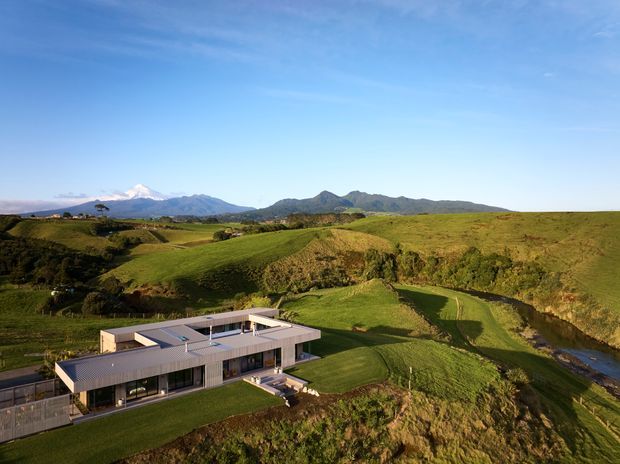 Aerial view of a contemporary single-storey residence set within expansive rolling green farmland. The house has a low-profile, rectilinear form with broad flat roof planes, extensive glazing, and a pale grey metal roof and facade elements. A winding stream runs through the landscaped rural site, with distant mountain ranges visible beyond.