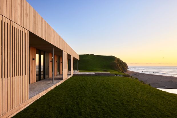 A contemporary coastal residence is shown from the exterior at sunset. The building has a long, low horizontal form with extensive glazing, a deep covered terrace, and vertical light timber cladding. It sits directly beside a grassy coastal headland overlooking a sandy beach and ocean. The warm timber facade contrasts with the dark green lawn, rocky landform, and pastel sky.