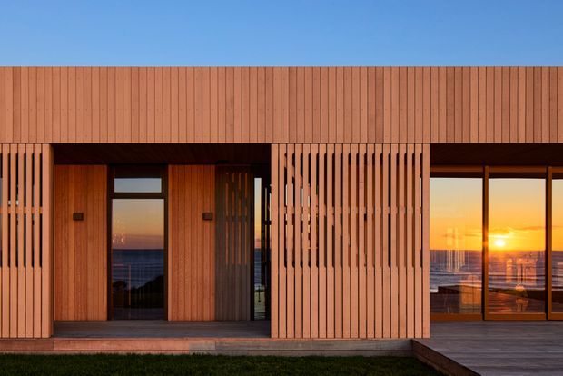 A contemporary timber-clad building facade at sunset, featuring vertical slatted screens, a recessed entry with glazed door, and expansive floor-to-ceiling glazing revealing a coastal or waterfront outlook. The warm timber envelope and strong horizontal and vertical geometry create a refined modern exterior.