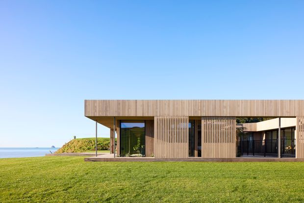 A low, minimalist contemporary house set beside open water, viewed from the lawn. The building has a strong horizontal flat-roofed form, extensive glazing, and warm vertical timber screening and cladding. Deep roof overhangs create shaded outdoor areas, while the expansive grass foreground and coastal setting emphasise the connection to the landscape.