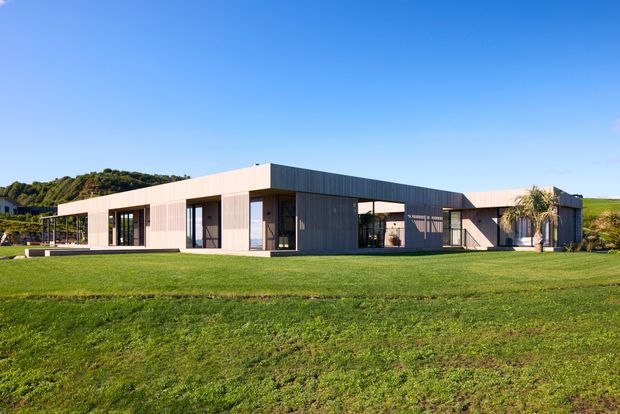Wide exterior view of a low, single-storey contemporary house set in an expansive lawn. The building has a long, rectilinear flat-roofed form with pale cladding, darker vertical-panel sections, extensive floor-to-ceiling glazing, and recessed covered outdoor areas. Rolling green terrain and a palm tree surround the home under a clear blue sky.