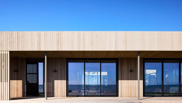 Front-facing exterior view of a contemporary low-rise building with a long horizontal facade clad in pale vertical timber battens. Large dark-framed glazed openings and sliding doors reveal bright interior spaces and expansive blue water or sky beyond. A covered terrace with timber decking runs along the facade, with strong sunlight creating alternating bands of light and shadow.