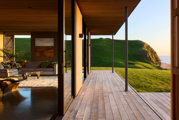 A warm, modern coastal home opening onto a broad timber deck with slender dark columns and expansive sliding glass doors. The interior living area is visible on the left, while the deck frames a dramatic green coastal headland and ocean beyond. The ceiling and deck use matching timber, creating a seamless indoor-outdoor connection.