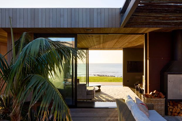 Interior view of a coastal living room opening directly onto a deck and oceanfront landscape. The room features extensive floor-to-ceiling glazing, warm timber-lined ceilings and flooring, dark structural framing, a large fireplace with exposed firewood, and relaxed lounge seating. A tropical palm partially obscures the left side of the room, while the ocean horizon is visible beyond the open glazing.
