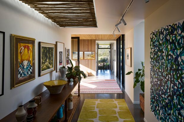 A long, bright residential hallway or gallery corridor with white walls, dark timber flooring, extensive framed artwork, a console table, indoor plants, and layered rugs. The corridor leads toward a timber-lined entry or living area with glazing and garden views. A distinctive exposed branch-like timber ceiling feature runs along the left side, while track lighting provides focused illumination. The space has an eclectic, art-filled character with strong natural materials and vivid textile accents.