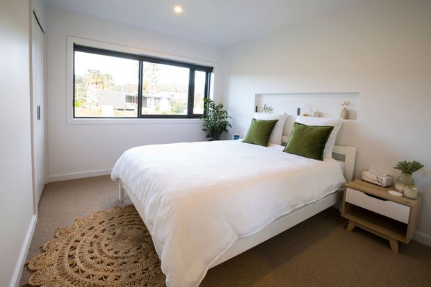 Bright, neatly styled bedroom with a low-profile white bed, white bedding, olive-green accent cushions, and a simple white floating bedside shelf. A large black-framed horizontal window provides daylight, while a woven round rug, potted plant, and small wooden bedside table add natural texture and warmth. The room has white walls, a plain white ceiling with recessed lighting, and neutral carpet.