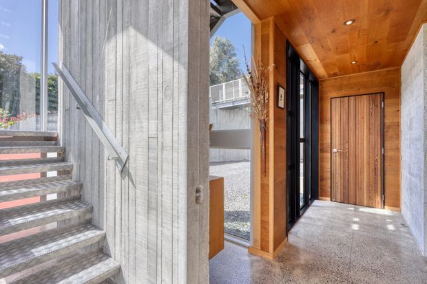A contemporary entryway viewed beside an exposed concrete stairwell. The space features a warm timber-lined ceiling and wall framing, a vertical timber front door, black-framed glazing, a polished grey terrazzo or concrete floor, and a contrasting raw concrete stair and wall surface. Daylight enters through large windows, creating a bright, refined indoor arrival space.