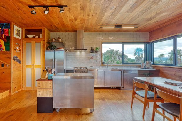 Warm, characterful kitchen with extensive timber wall and ceiling lining, stainless-steel island and appliances, white subway-tile splashback, timber flooring, and large corner windows overlooking greenery and palms. The room includes a dining table, open shelving, and colourful eclectic artwork and accessories.