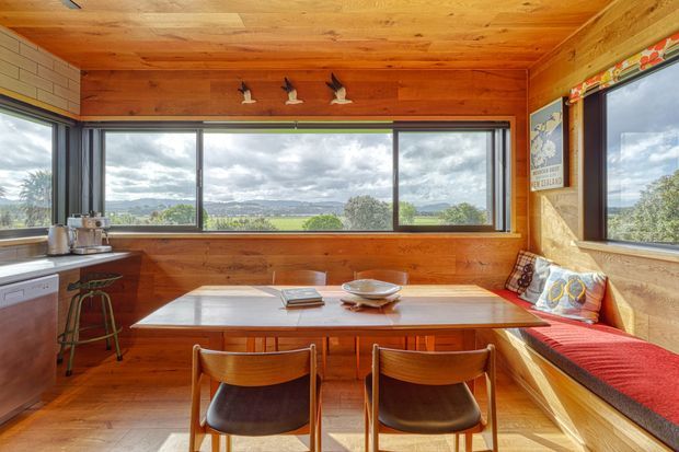 A warm, compact dining nook with a built-in timber bench beneath expansive corner windows. A wooden dining table with matching chairs occupies the centre, while a small kitchen work area with stools is visible at left. Timber lines the walls and ceiling, and the windows provide expansive views across a green rural landscape and hills. Decorative bird figures, cushions, and a red bench cushion add character.