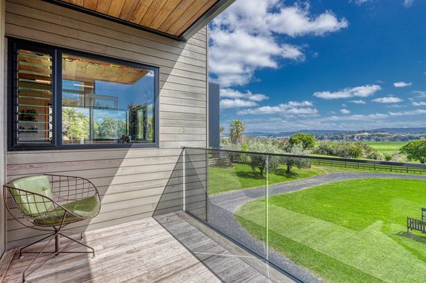 A modern outdoor balcony or terrace attached to a contemporary home, featuring horizontal grey weatherboard cladding, timber soffit and decking, frameless glass balustrading, and a dark-framed window reflecting the interior. The terrace overlooks a manicured lawn, driveway, rural fields, and distant hills beneath a bright blue sky.