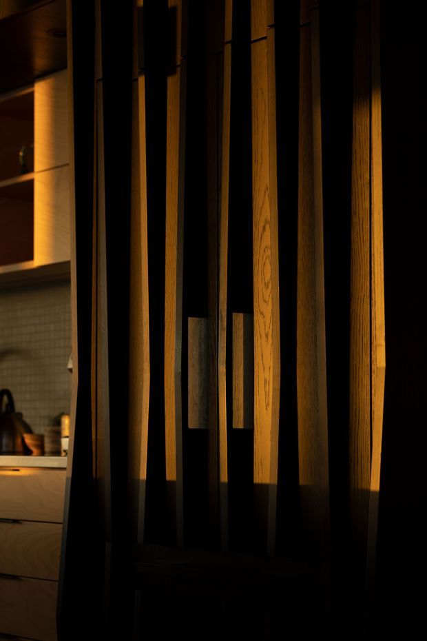 A moody close-up of a dark timber slatted screen or partition with warm concealed lighting. The vertical timber battens create a strong architectural feature, while a partially visible kitchen with timber cabinetry, shelving, tiled splashback and a kettle appears behind the screen at left.