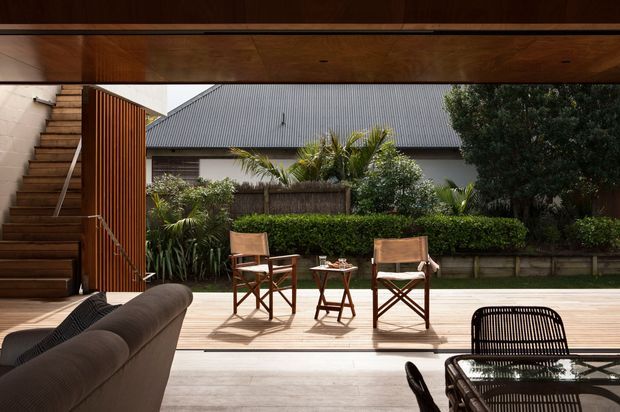 A view from inside a contemporary home onto a timber-decked outdoor entertaining area. The scene includes a broad timber-framed opening, internal timber stair and vertical timber screen, outdoor director-style chairs and a small table, a partial sofa in the foreground, and a landscaped garden beyond. A neighbouring or background building with a corrugated metal roof is visible past the garden.