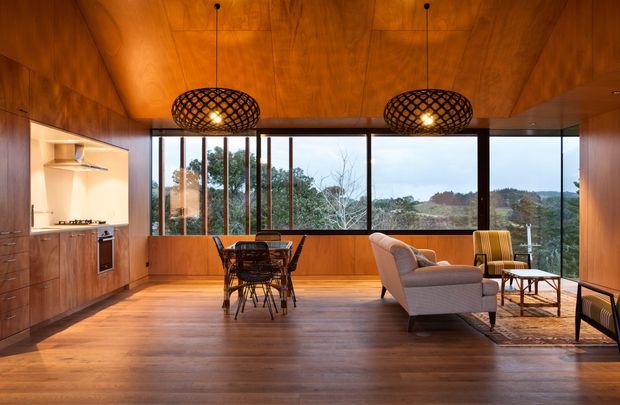 A warm, open-plan living and dining area with a compact kitchen along the left wall. Rich timber lining covers the ceiling, walls, cabinetry, and flooring, while large floor-to-ceiling windows provide views of a wooded landscape. The room contains a dining table and chairs, a sofa, lounge chairs, a coffee table, and two woven pendant lights. The design has a strong mid-century character with extensive timber finishes and simple modernist glazing.