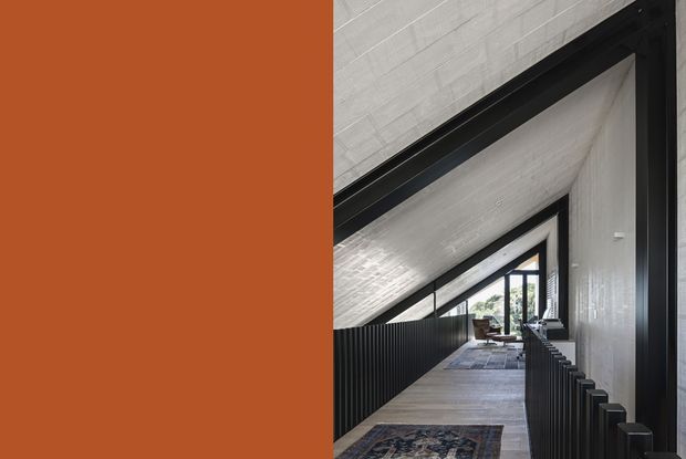 A narrow, dramatic interior passage or landing beneath a steeply sloping timber-lined ceiling. Dark black metal balustrades frame the walkway, with large black-framed glazing at the far end and a glimpse of seating beyond. The palette is restrained, combining warm terracotta, pale timber, grey, and black architectural elements.