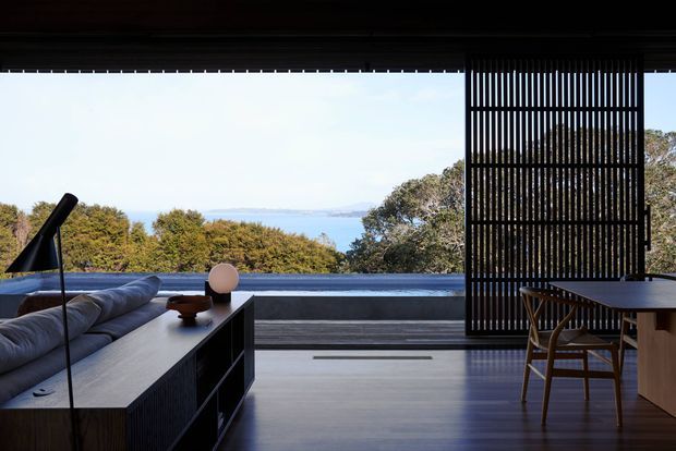 A refined open-plan living and dining interior framed by expansive glazing, with a panoramic coastal or waterfront outlook beyond. The room features a dark timber-toned built-in media/storage unit, low contemporary seating, a dining table with timber chairs, a black sculptural floor lamp, and a vertical slatted privacy screen. The polished dark floor and restrained furnishings create a calm, sophisticated atmosphere with strong indoor-outdoor connection.