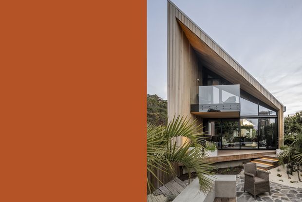 Exterior architectural photograph of a contemporary multi-level home with a dramatic steeply sloping roof, extensive floor-to-ceiling glazing, vertical timber cladding, and a projecting upper balcony with a clear glass balustrade. The house is set within landscaped coastal-style planting and a paved outdoor entertaining area. A large terracotta-coloured vertical panel occupies the left side of the image.
