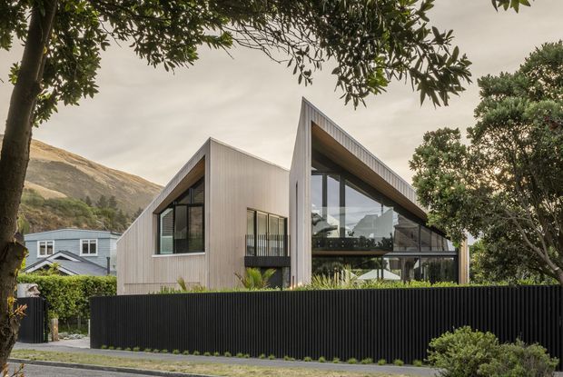 Exterior view of a contemporary single-storey house with two steeply pitched gabled roof forms, light vertical cladding, extensive floor-to-ceiling glazing, and a sheltered glazed living area. The home is screened by a tall black vertical-slat fence and landscaped planting, with a rugged hillside visible behind it.