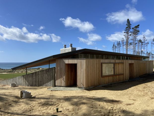 Single-storey timber-clad coastal building viewed from outside, with a low-pitched asymmetrical roof, broad roof overhang, small chimney vents, and simple timber-framed openings. The structure sits beside a sandy, windswept site with ocean visible beyond and tall coastal trees in the background.