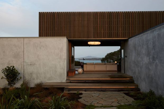 A low, modern coastal home entrance framed by smooth concrete walls and a tall vertical timber privacy screen. Broad timber steps lead through a covered passage into a sheltered outdoor sitting area, with a glimpse of blue water and distant hills beyond. Native-style planting and irregular stone paving soften the minimalist architecture.