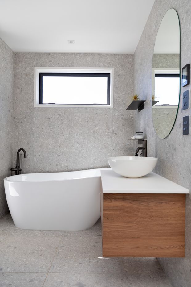 A compact contemporary bathroom with a freestanding white bathtub, wall-mounted timber vanity with a white benchtop, white vessel basin, matte black tapware, oval wall mirror, and a small horizontal window. Large-format grey stone-look tiles cover the walls and floor.