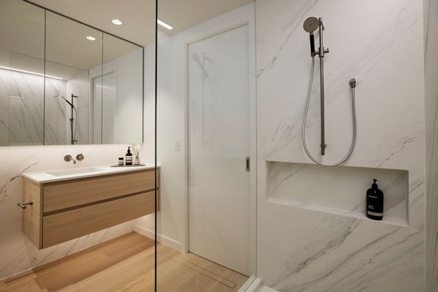 Contemporary minimalist bathroom with a frameless glass shower enclosure, large white marble-look wall tiles with grey veining, a recessed shower niche, wall-mounted brushed metal shower fittings, a floating light-wood vanity, integrated basin, and large mirrored cabinetry. The palette is light, warm, and refined.