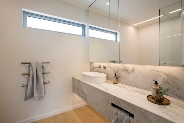 Contemporary bathroom with a floating stone vanity, integrated basin, large mirrored wall cabinets, frameless glass shower enclosure, horizontal clerestory windows, and a heated towel rail. The space uses warm timber flooring, pale walls, and veined light stone for a clean, refined appearance.