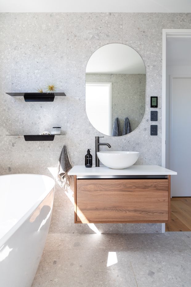 Bright contemporary bathroom with pale grey stone-look wall and floor surfaces, a freestanding white bathtub, floating timber vanity with white vessel basin, matte dark tapware, oval arch-top mirror, and minimal open shelving. A doorway and timber flooring are visible at right.