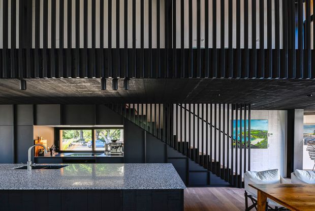 Interior view of a dark, contemporary open-plan home showing a kitchen island with a speckled stone benchtop, black cabinetry, and integrated appliances. A dramatic open stairway with closely spaced black vertical balusters rises through the centre, beneath a dark timber-lined upper floor or mezzanine. The dining area and living seating are visible to the right, with timber flooring and large windows providing views and daylight.