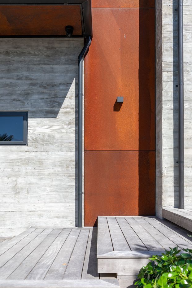 Close-up exterior view of a contemporary building entry or facade. The composition features weathered grey timber cladding, a tall rust-coloured metal feature panel with visible fixings, dark metal framing and downpipes, a small window, and a broad timber deck with a raised step or bench-like platform. Strong sunlight creates crisp shadows, with limited planting visible at the edge.