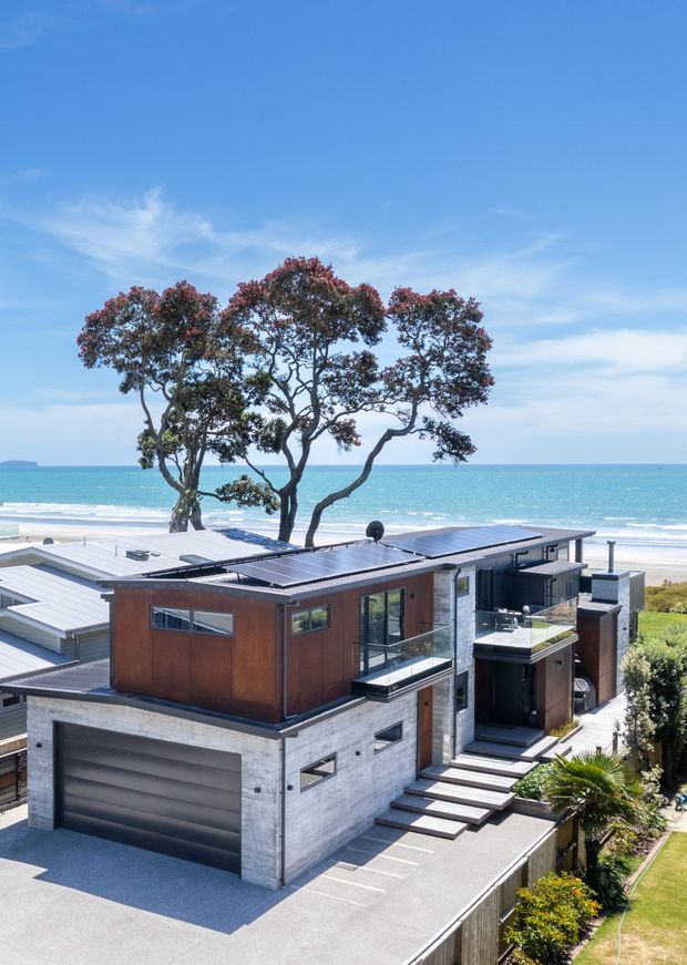 A contemporary multi-level coastal house photographed from above, featuring flat rooflines, concrete and dark timber-toned cladding, extensive glazing, rooftop solar panels, balconies, and a large integrated garage. The property overlooks a sandy beach and turquoise ocean, with mature trees framing the view.