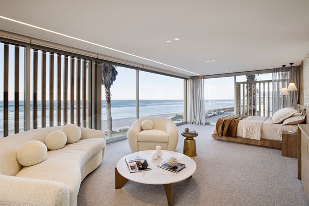 A bright, open-plan coastal living room and bedroom suite with floor-to-ceiling glazing overlooking the ocean. The room features a curved cream sectional sofa, rounded armchair, circular coffee table, neutral carpet, linen bedding, timber bedside cabinetry, sheer curtains, and vertical timber screening elements. The interior has a soft, refined contemporary feel with abundant natural light and a direct beach outlook.