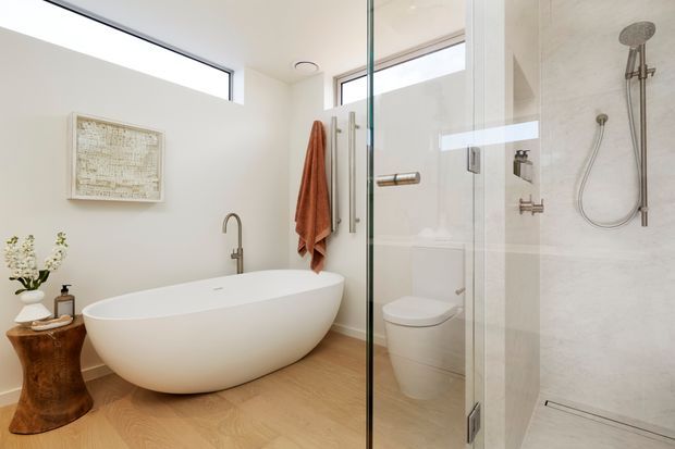 Bright contemporary bathroom with a freestanding white oval bathtub, wall-mounted bath filler, frameless glass shower enclosure, rainfall and handheld shower fittings, and a concealed toilet. Pale timber flooring, white walls, a large horizontal window, and warm wood accents create an airy, refined space.