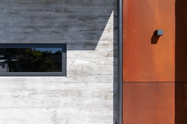 Close-up architectural exterior detail showing a pale weathered timber-clad wall with a dark-framed horizontal window beside a rust-orange panelled facade. A small projecting exterior light or fixture is mounted on the orange wall, with strong sunlight creating sharp shadows.