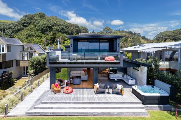 Contemporary multi-level residential home set into a leafy hillside, viewed from the rear. The dark-clad house features broad glazing, a glass-balustraded upper terrace, expansive timber decking and stepped outdoor entertaining areas. An open-plan living space is visible on the lower level, with outdoor seating, colourful beanbags and a spa pool. Neighbouring homes and dense greenery surround the property under a bright blue sky.