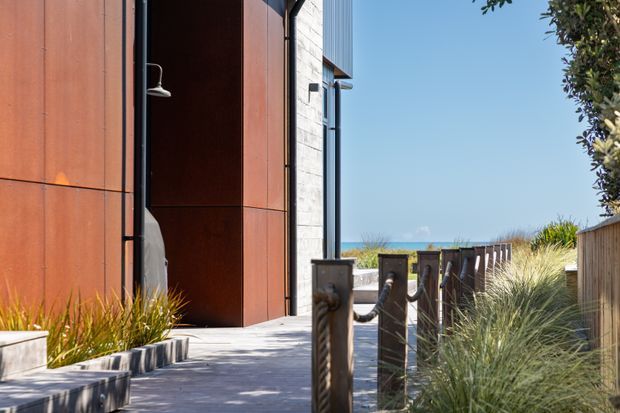 Exterior architectural view of a contemporary coastal building, featuring a tall rust-toned metal-clad wall, exposed pale masonry, large vertical glazing, and a recessed entry. A paved walkway with timber-and-metal post fencing leads toward dune grasses and a visible ocean horizon.
