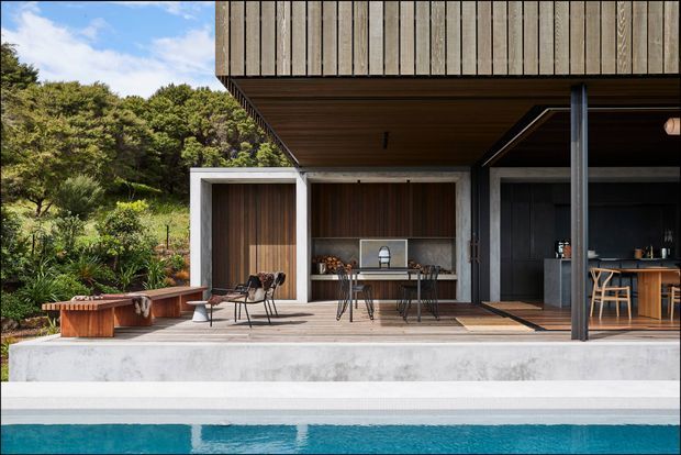 A contemporary open-plan outdoor entertaining area beside a swimming pool, set against dense greenery. The space features a broad concrete terrace and step, dark timber-lined ceilings and walls, vertical timber cladding above, a dining setting, an outdoor kitchen or bar area, a built-in bench, and slender dark metal posts. Large openings connect the terrace to the sheltered interior, creating a refined indoor-outdoor living arrangement.