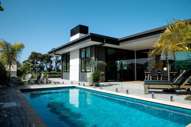 A modern single-storey residence viewed from the pool area, featuring a crisp flat-roof form, white masonry walls, dark trim, extensive floor-to-ceiling glazing, and a spacious turquoise swimming pool with a paved surround. The covered outdoor living area contains dining furniture and loungers, with palms and established landscaping around the property.