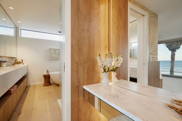 Warm, refined bathroom interior viewed from an adjacent vanity or dressing area. The space features a long timber vanity with a pale stone benchtop, full-height timber wall panelling, a freestanding white bathtub, light timber flooring, mirrored surfaces, and a coastal ocean view through the glazing. A sculptural carved feature panel and neutral finishes add decorative detail.