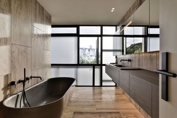 Contemporary bathroom featuring a freestanding dark bathtub, wall-mounted black tapware, a long floating timber vanity with integrated basin, and a full-height mirror above the vanity. Large black-framed windows and glazed doors bring in abundant natural light, while timber-look wall and floor finishes create a warm, refined atmosphere.