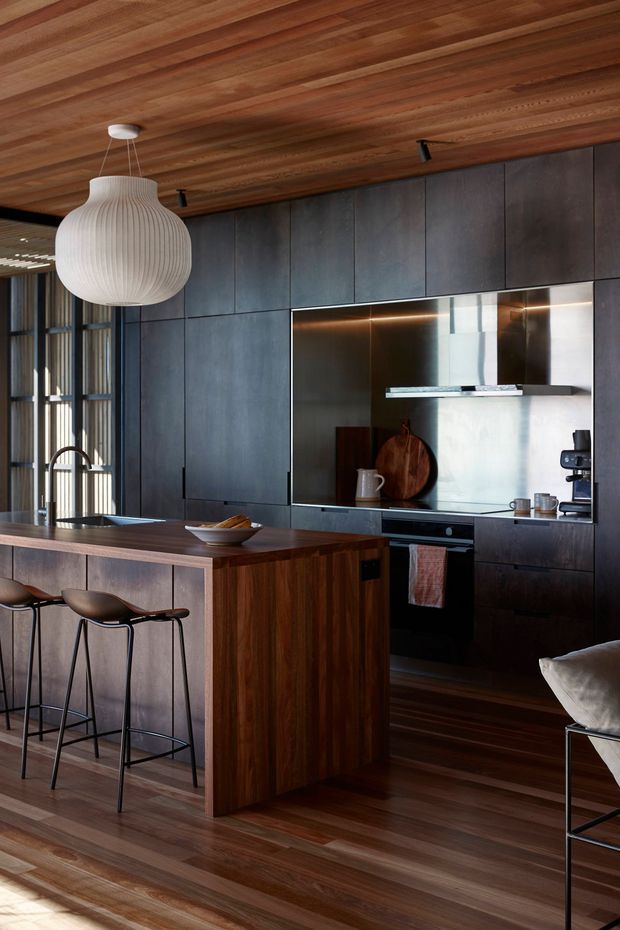 A sophisticated open-plan kitchen with dark charcoal built-in cabinetry, a timber island and matching timber floor and ceiling. The kitchen features an integrated cooktop and oven, stainless steel rangehood, under-cabinet lighting, a dark stone-like benchtop, and sculptural pendant lighting. Bar stools line the island, while a partial lounge chair is visible at the right edge. Tall glazed openings with vertical timber screening bring in natural light.