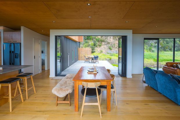 Open-plan contemporary living and dining area with a warm timber ceiling and light timber flooring. A substantial timber dining table sits centrally between the kitchen and lounge, with large glazed sliding doors opening to an outdoor dining deck and landscaped setting. Blue upholstered seating is visible to the right, while the kitchen with island bench and stainless-steel refrigerator is visible to the left.