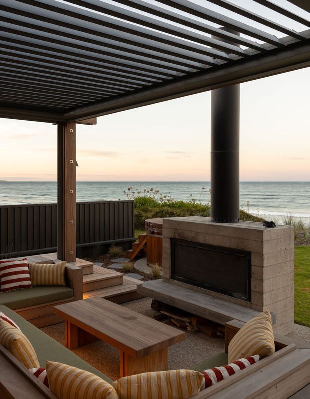 A covered outdoor lounge overlooking the ocean, with built-in timber seating, striped cushions, a low timber coffee table, dark privacy screening, and a substantial masonry fireplace with a black chimney. A slatted pergola roof filters the warm coastal light, creating a sheltered beachfront entertaining area.