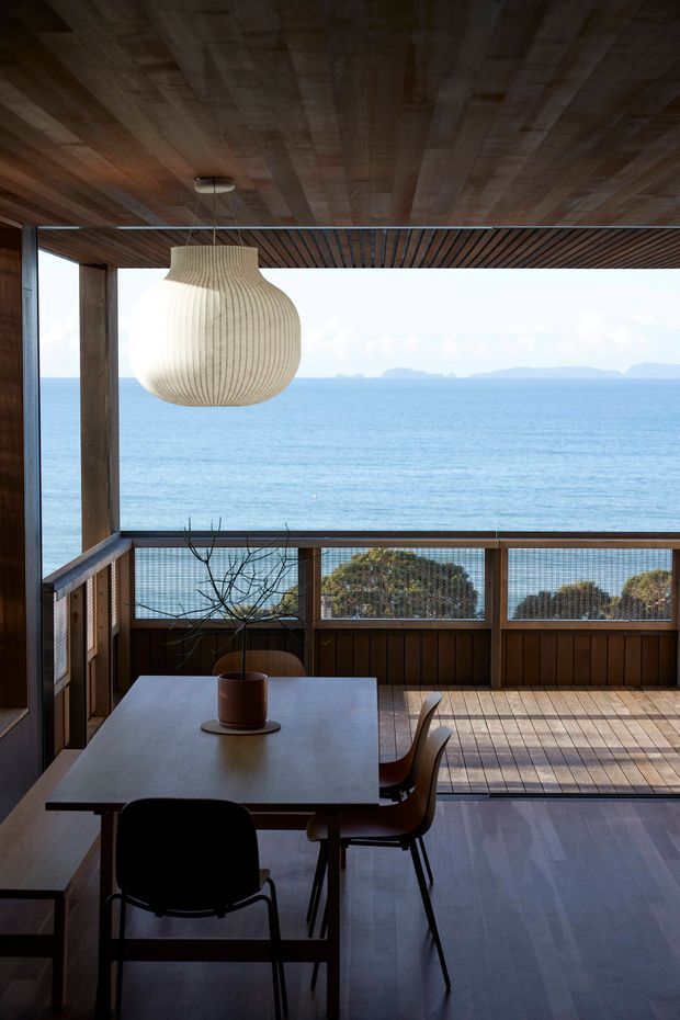 A compact dining area in a timber-lined coastal home, opening onto a broad ocean view through extensive glazing. The room features a rectangular dining table with dark chairs, a sculptural woven pendant light, timber ceiling and wall cladding, and a timber deck or balcony beyond the dining space. The atmosphere is calm, warm, and strongly connected to the sea.