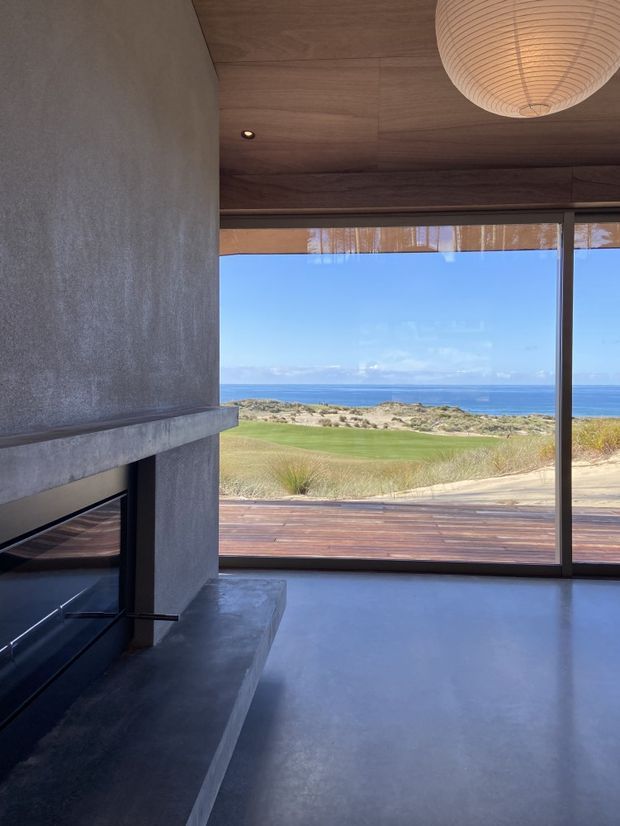 A minimalist living room with a dark concrete fireplace surround and hearth, smooth grey polished flooring, and a warm timber-lined ceiling. Full-height glazing opens to a timber deck and expansive coastal ocean views, while a large woven pendant light adds a soft natural accent.